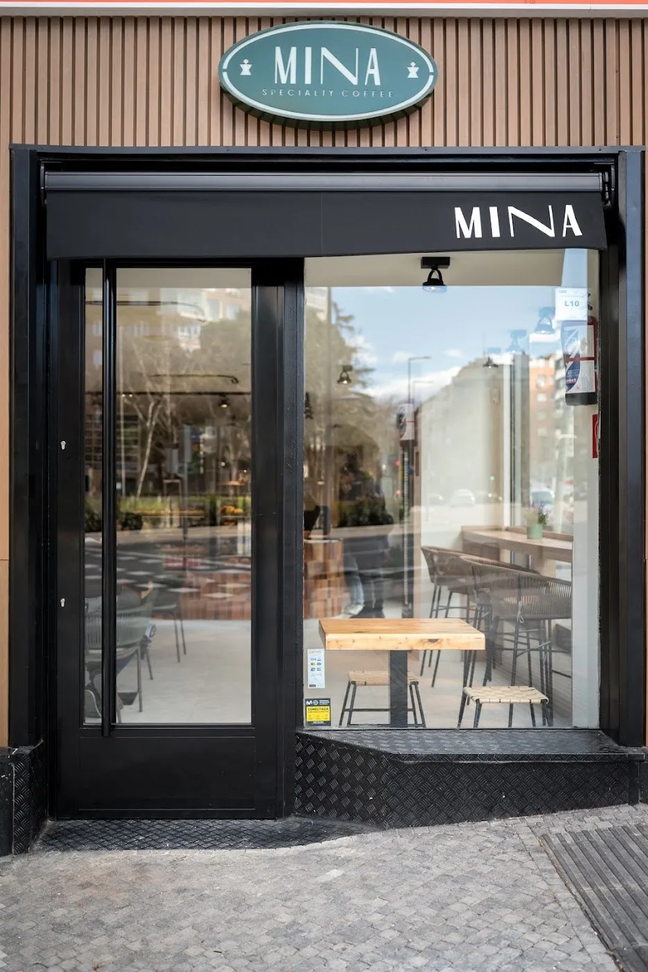 Fachada negra con la placa ovalada verde de Mina sobre lamas de madera y el toldo con su nombre