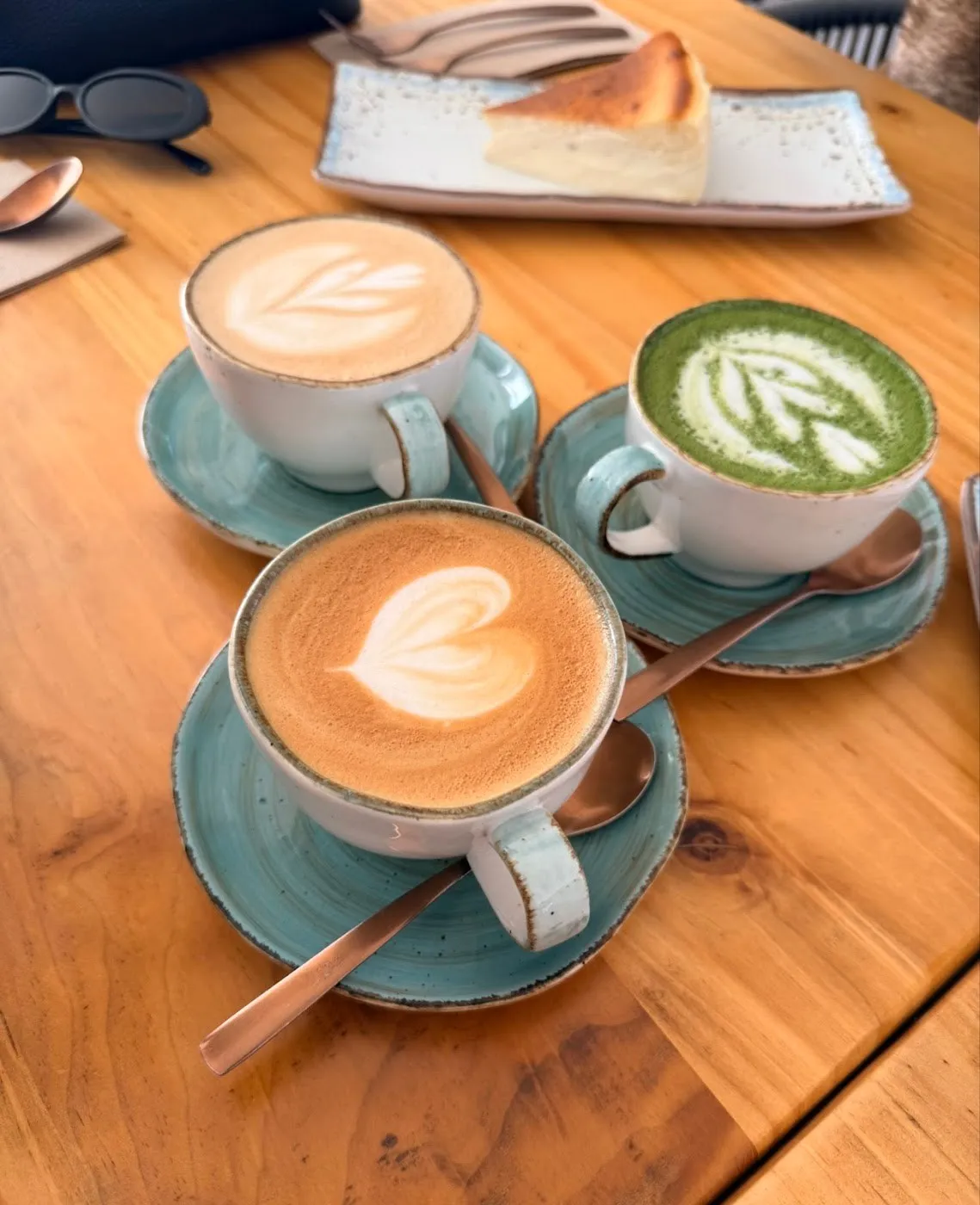 Tres cafés, uno de ellos matcha, en tazas turquesa y una porción de tarta de queso